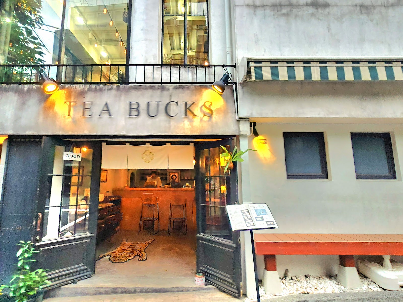 夕方の店先。コンクリートの鴨居に大文字で TEA BUCKS とあり、二つの暖色のスポットが当たっている。黒い鉄の扉が開き、金色の紋の入った白い暖簾の奥にテラコッタ色の壁のカウンターと二脚のスツール。床にトラ柄のラグ、脇に鉢植え。